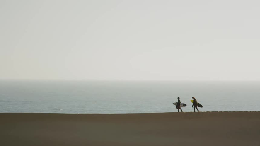 Two people walk along the beach carrying surfboards