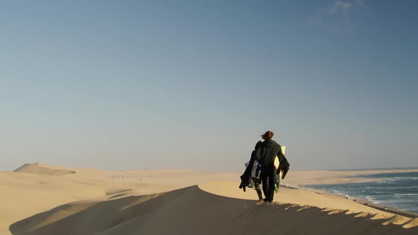 Two surfers walk along a sand dune near the coastline
