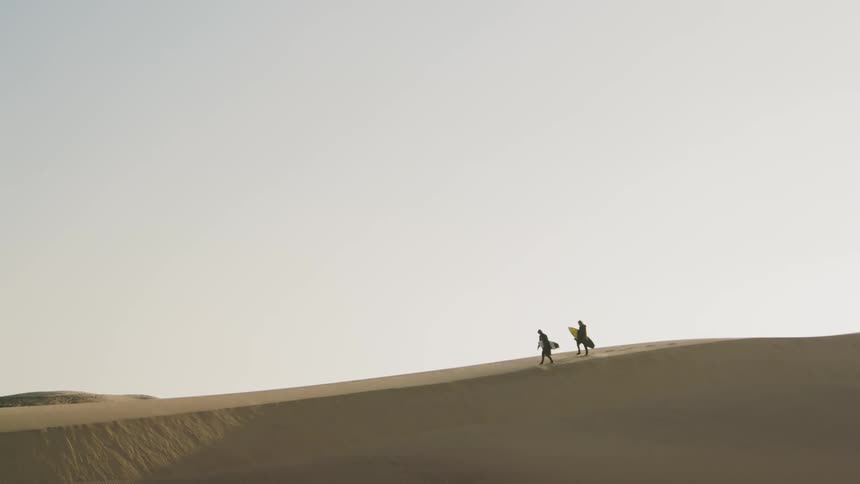 Men holding surfboards walk on the sand dunes in the desert