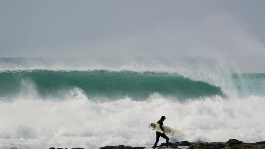 Man with surfboard stands on the rocky beach next to big crashing ocean waves
