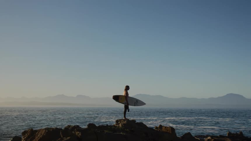 A woman holds a surfboard standing along coastal rocks