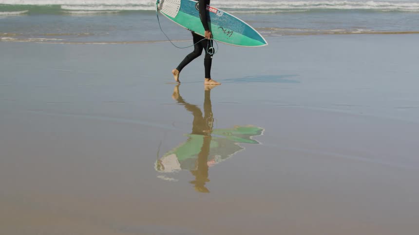 A person walks across the shoreline holding a surfboard