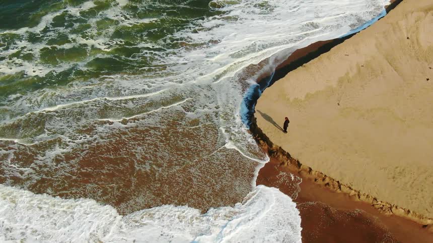 A person walks to the edge of a cliff to look at the water rushing in to shore
