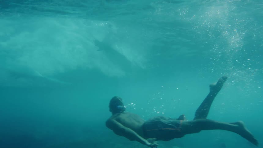 A white male swims underneath a surfer riding a wave in the ocean