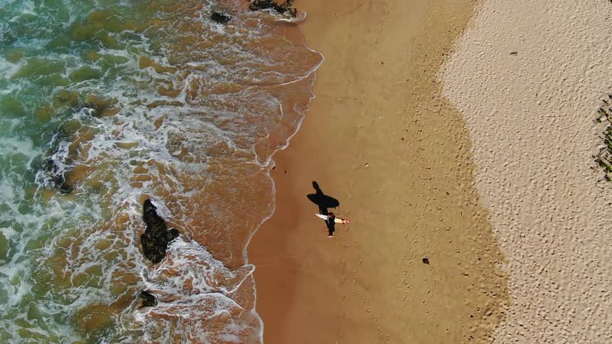 Man holding a surfboard stands on the beach and watches the wavy sea