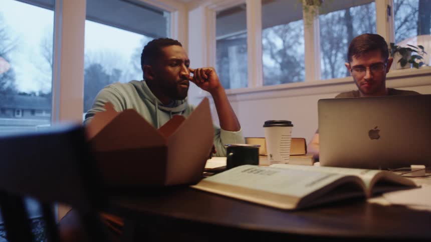 Two men sit at a table with a laptop and books and papers