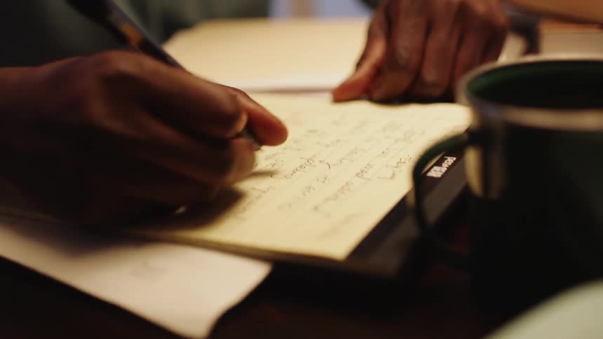 Man writes on a piece of paper with a pen as a coffee mug sits nearby