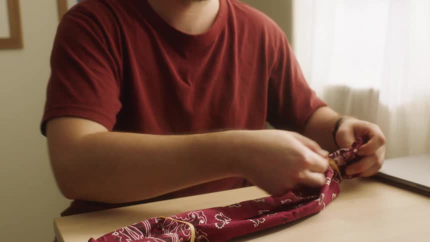 A man sits at a table and places hair ties on a bandana to make a face mask
