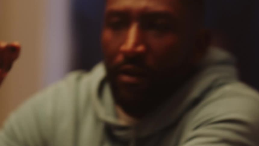 A black man points at a laptop screen with a pen and then sits back and puts on his hood