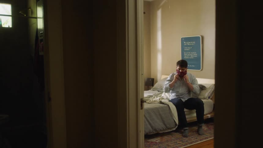 A man sits on his bed and puts on a protective mask and then walks out the door