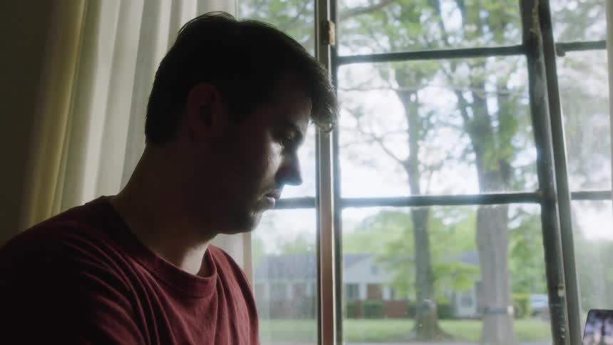 A man is looking at a laptop while sitting by a window