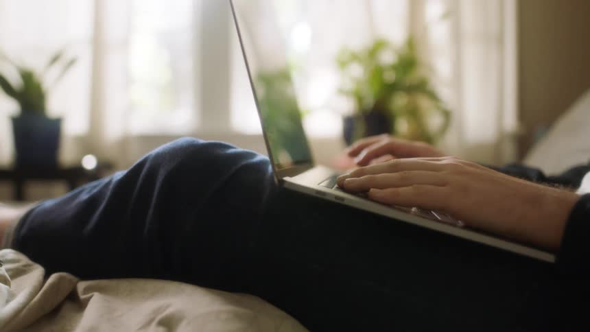 Person reclines on bed typing on laptop held in their lap