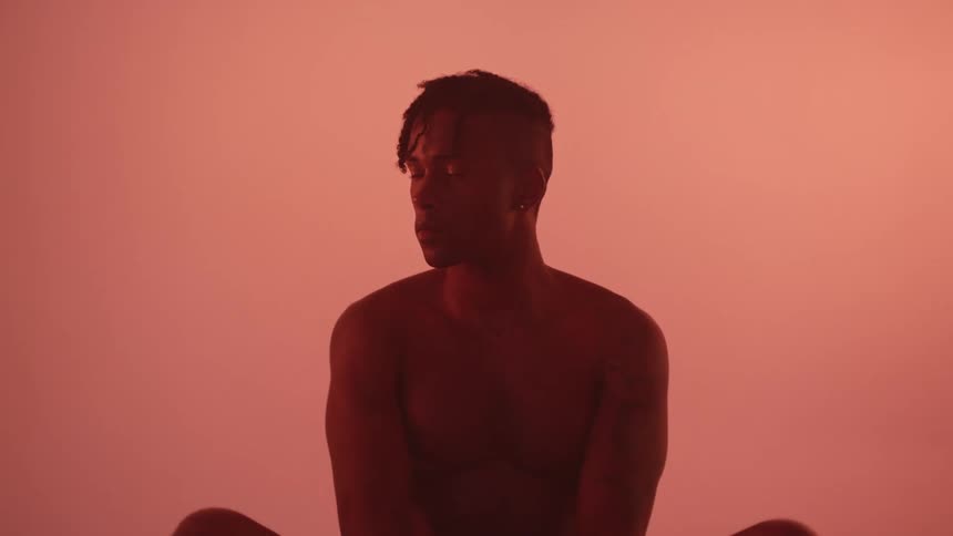 A man sits naked with his legs crossed in front of a pink wall