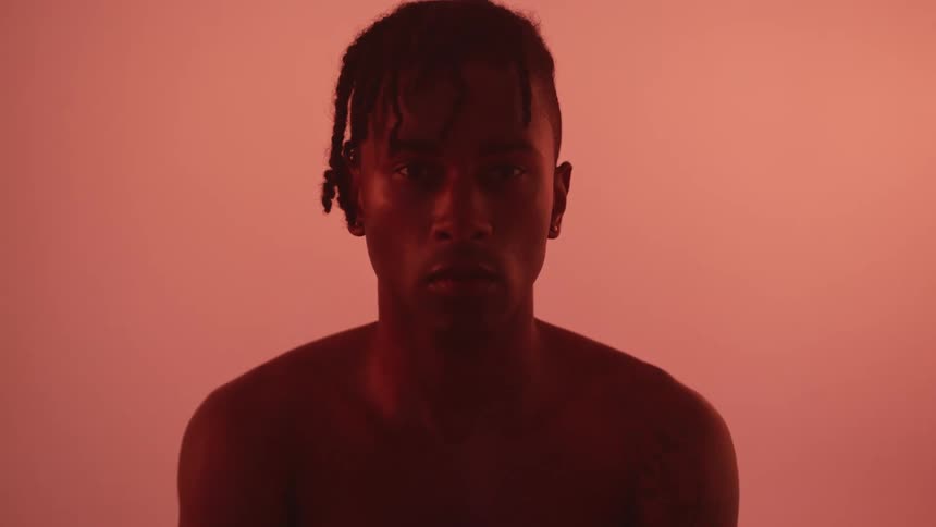 A naked man sits cross legged staring straight ahead with a pink background