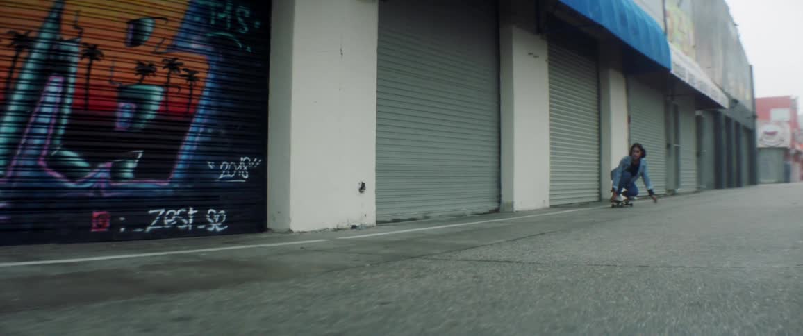 A female skateboarder rides down a city sidewalk in front of close storefronts