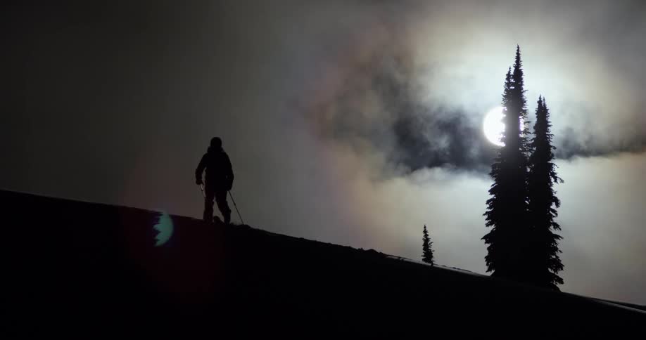 The silhouette of a skier on the ridge of a slope with trees behind and a full moon in a cloudy sky