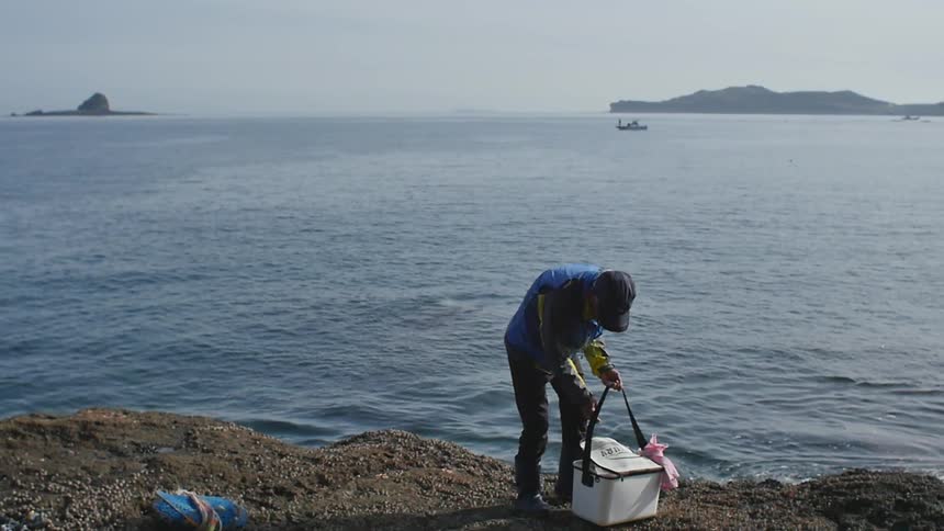 A man stands near a body of water and looks through a lunchbox