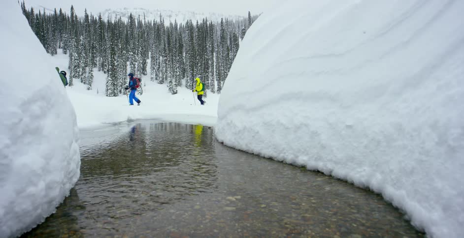 Three people walk across snow covered terrain by a lake and forest