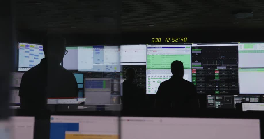 Three men stand inside a control room watching multiple monitors
