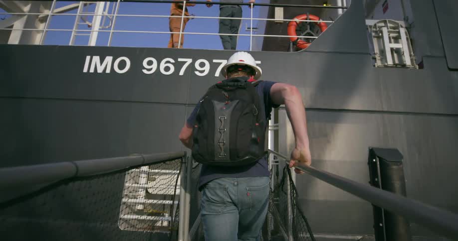 A man with a backpack and helmet walks up a gangway to enter a ship