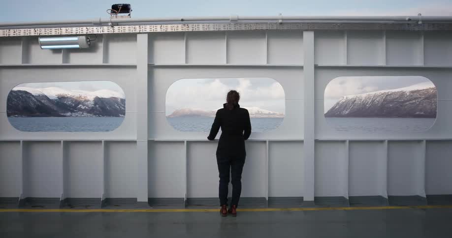 A woman stands on an open air boat and looks out a window at the water