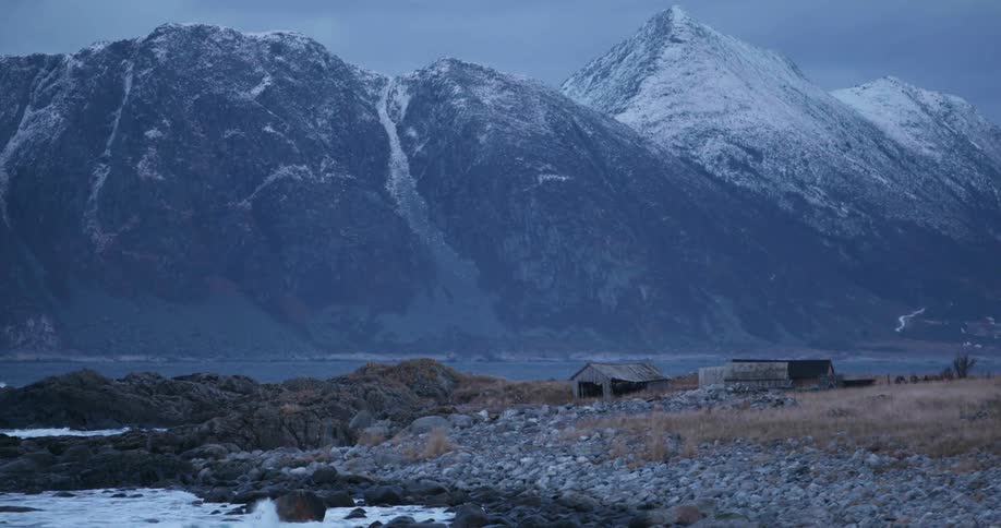 Abandoned wooden buildings on a rocky beach beneath a snow capped mountain