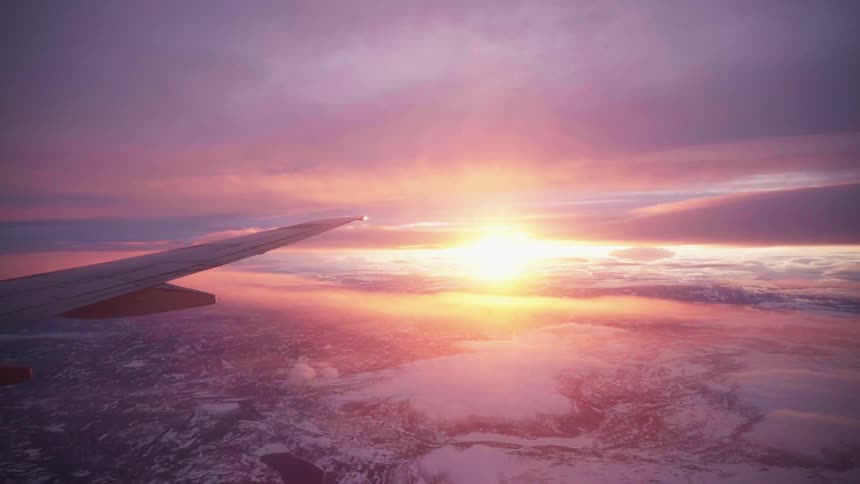 View of an airplane wing as it flies through the air and the landscape below as the sun sets