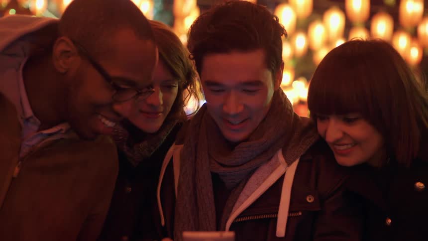 A man holds his phone and scrolls through it as his three friends look at it and smile
