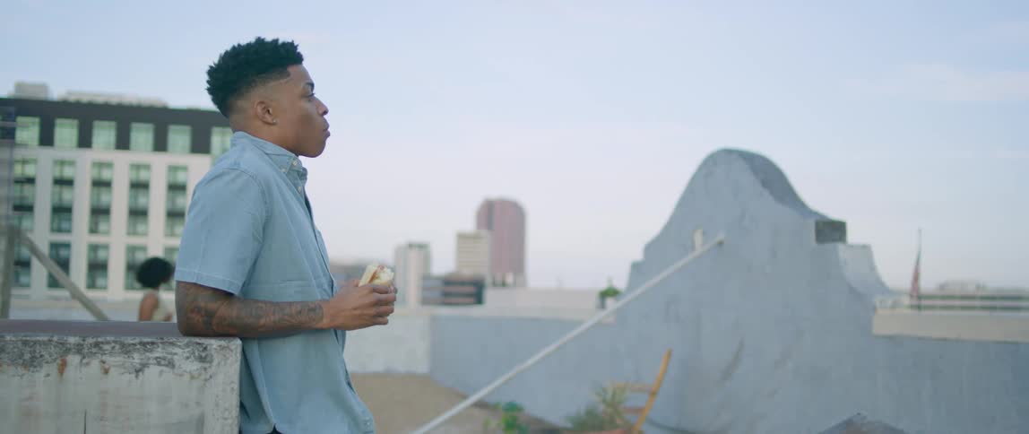 A black man eating a sandwich on a rooftop notices an attractive black woman walking nearby