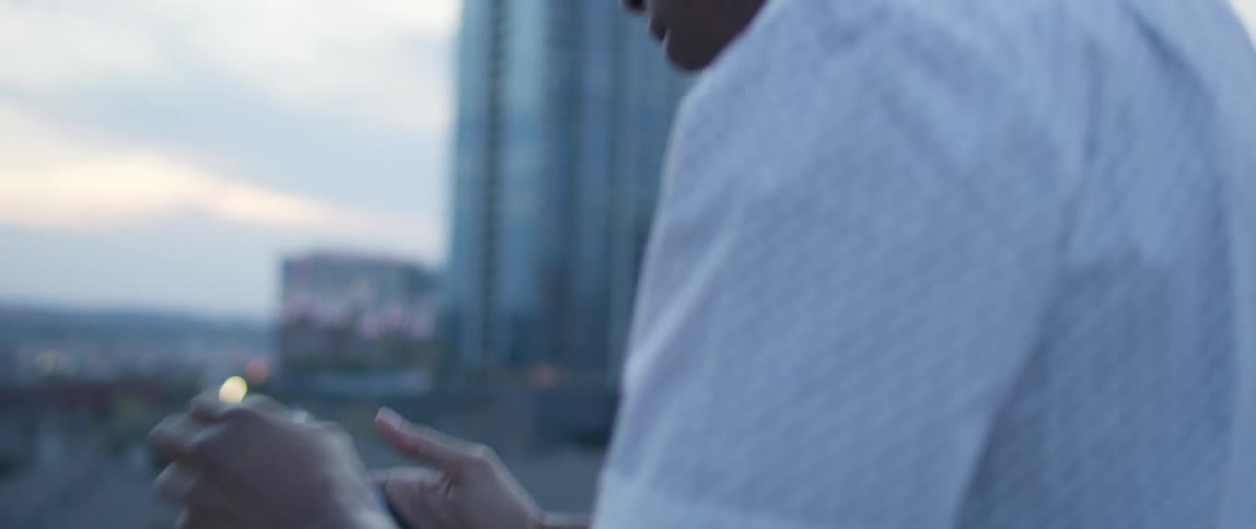 Male leans against the ledge of the balcony in a highrise building as he cues the music on his cell phone and listens through wireless earbuds while nodding his head to the beat