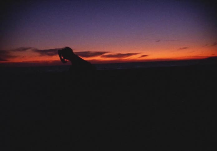 A woman stands outdoors and dances as the sun sets