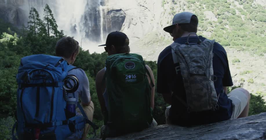 Three people with backpacks are sitting near a waterfall
