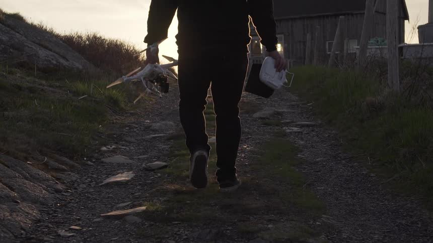 Man walking on a dirt path carries a drone at sunset