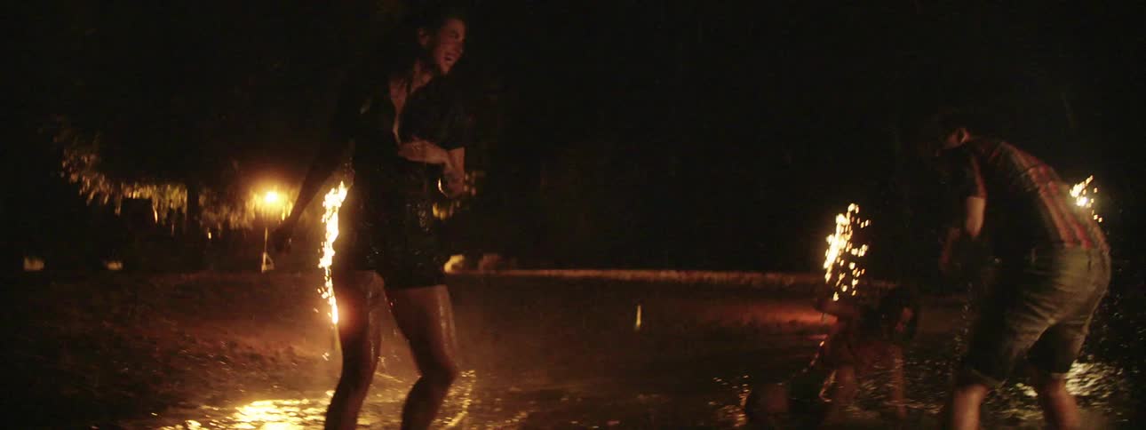 Two women and a man splash in water at a beach while fireworks go off nearby