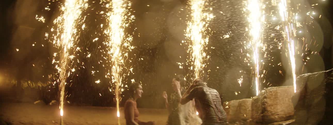 Friends splash water on each other as they stand in a field surrounded with sparking fireworks