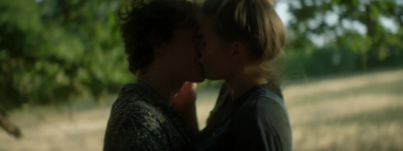 Teenage boy and girl kiss while they stand in a field near the forest