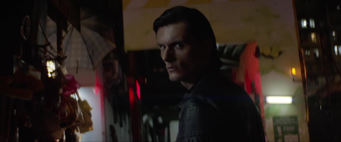 In narrow cluttered hallway man stares over shoulder with glare