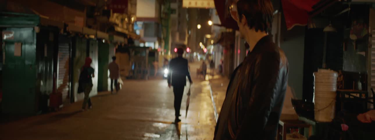 A man walks out to a city street at night and looks around the block