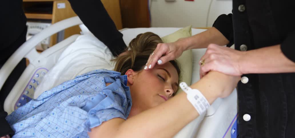 A teenager lies in a hospital bed with her eyes closed as her mother holds her hand and strokes her hair and her dad rubs her back
