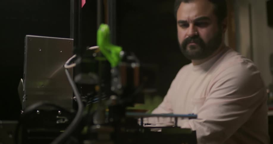 A man sits in an office at a desk waiting for a 3d printer to finish printing
