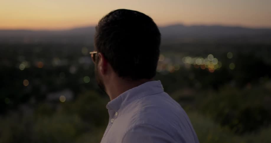 Man with beard and sunglasses stands on a hill and looks down at the distant town at dusk