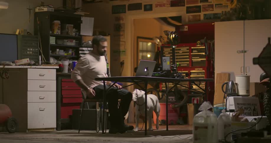 A man walks in to a room with his dog and sits down at his laptop