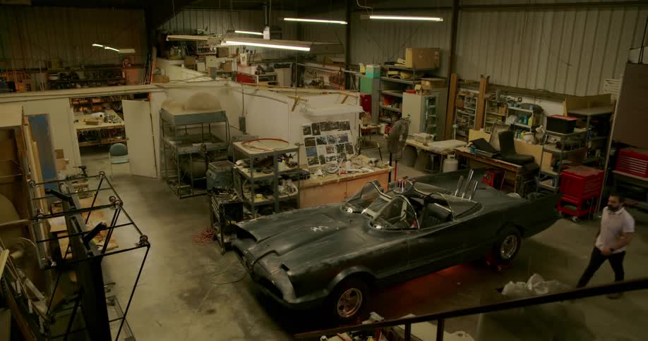 The lights come on in a large garage workshop and a man walks past a car towards the office area