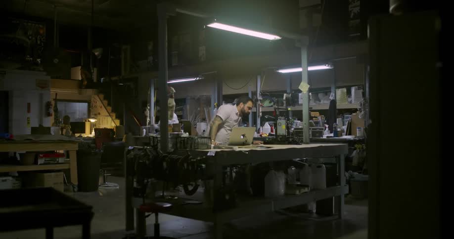 A man standing before a workbench in a dimly lit workshop using a laptop