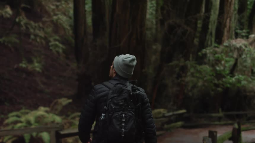 Male carrying a backpack with a tripod in the side pocket walks along a man made hiking trail lined by a wooden rail fence through the forest