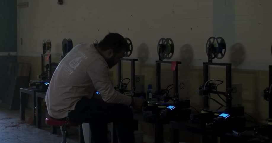 A man slides on a chair checking a row of 3d printers