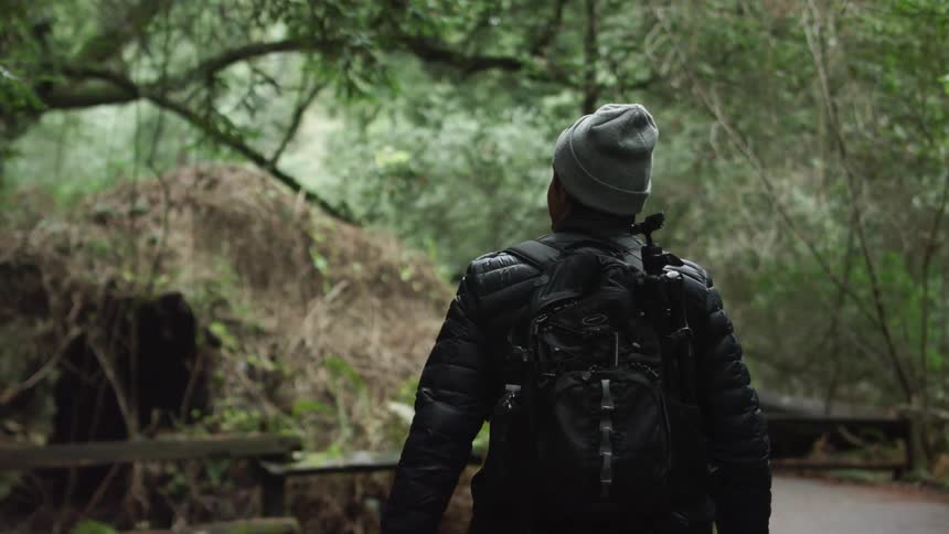 A man wearing a jacket and backpack looks around as he walks a paved trail through the trees