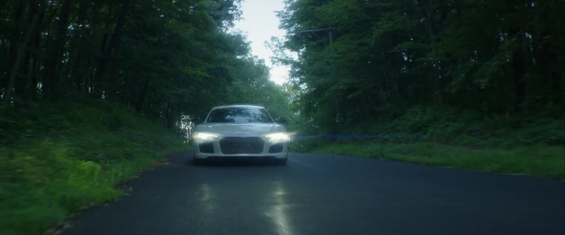 A white audi sports car driving on a road in a forest with its lights on
