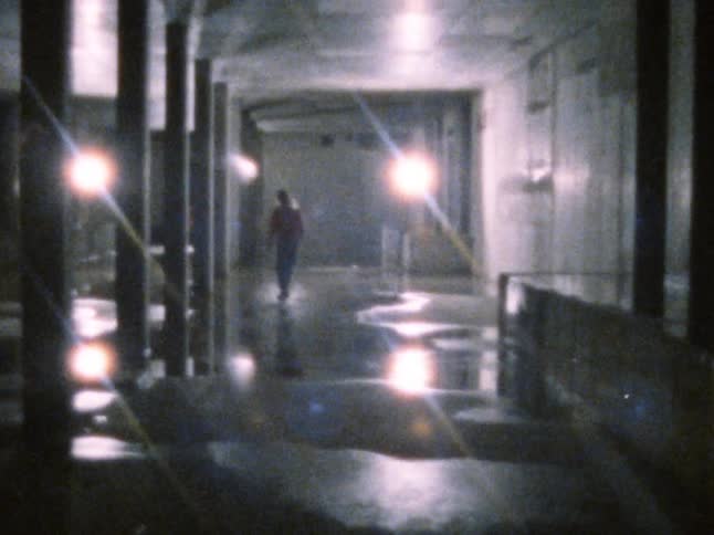 A friend watches as another person skateboards across a floor covered in water inside a dark building