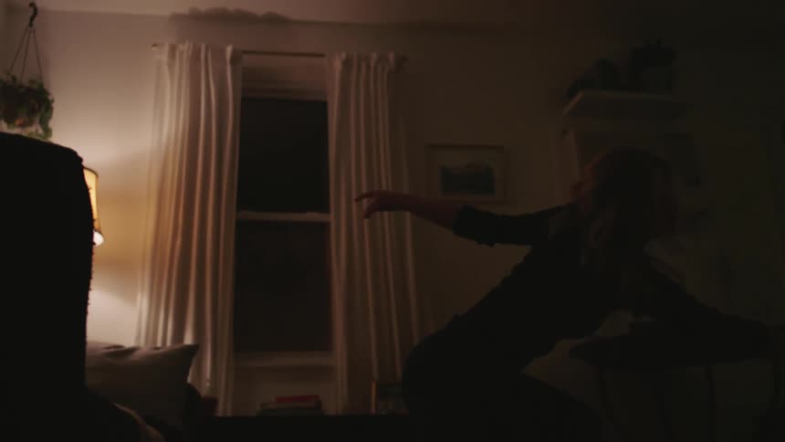 A blonde white woman dances in her living room at night